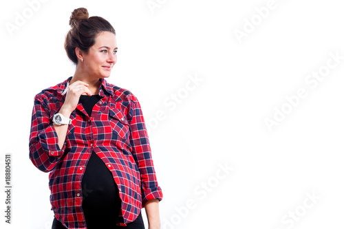 Young dark-haired pregnant woman in black dress and plaid shirt smiling and posing on isolated white background