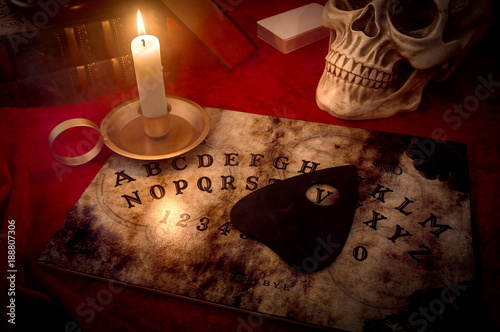 A witches den, fortune teller and black magic concept with a talking spirit board lit by a candle surrounded by a deck of cards, three books and a skull engulfed in mysterious and murky smoke