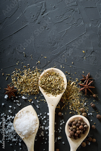  Wooden spoon with Italian seasoning-dried oregano with thyme, basil and vegetables.Oregano in a wooden spoon on a rocky concrete dark black background with a place for text.Top view.salt crystals pep