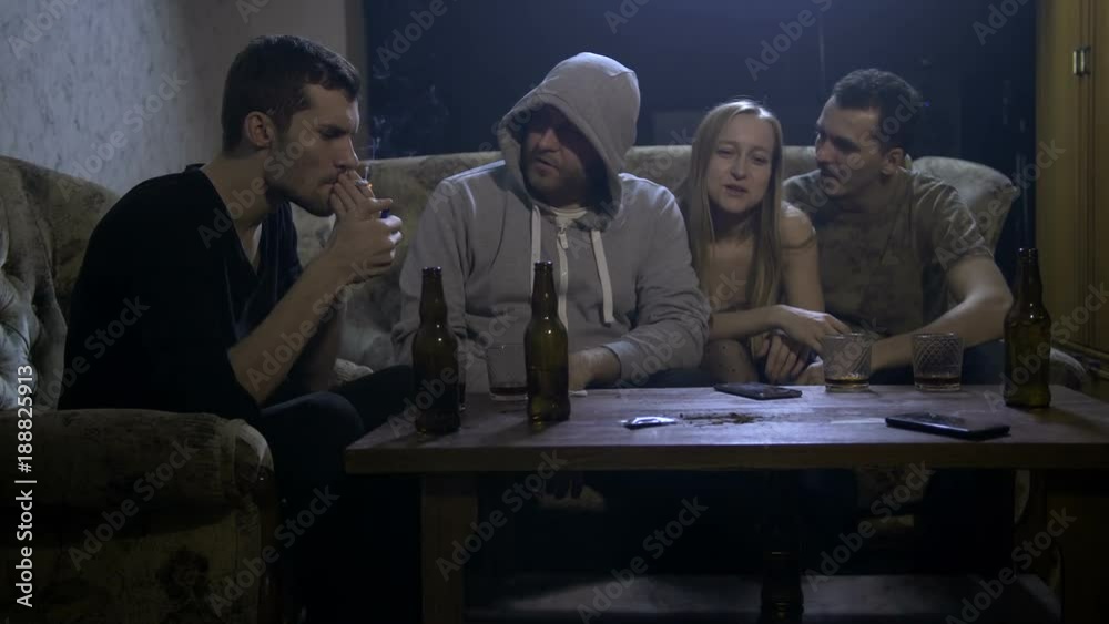 Group of four young drug addicts smoking cannabis at home at night on the sofa. One female and males passing marijuana joint to each other. Social issues, drug abuse and drug addiction concept