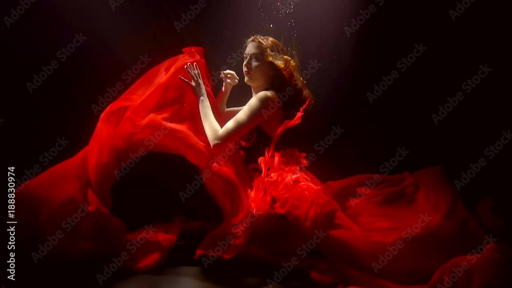 Attractive woman with bright red hair wearing bright red dress doing art underwater in a dark.