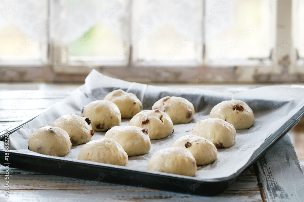 Raw unbaked buns. Ready to bake homemade Easter traditional hot cross buns on oven tray with baking paper over white wooden table with window at background. Natural day light. Rustic style. Raw unbaked buns. Ready to bake homemade Easter traditional hot cross buns on oven tray with baking paper over white wooden table with window at background. Natural day light. Rustic style.