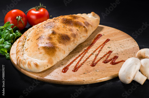 Fotografi Delicious Pizza calzone with basil leaves on wooden board on dark background