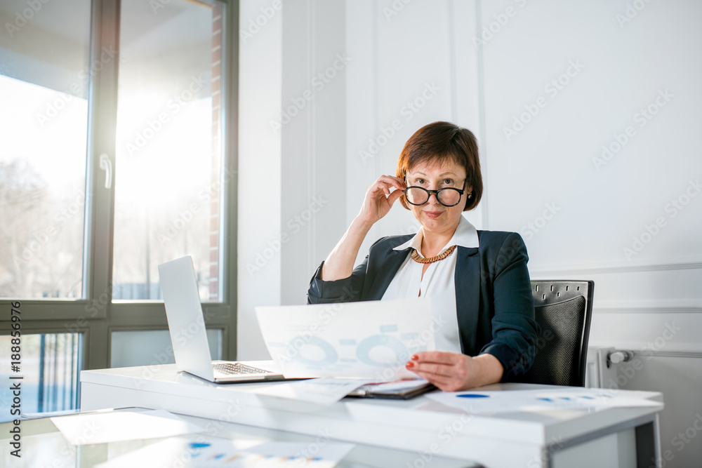 Portrait of an elegant senior businesswoman dressed in the suit working with laptop and documents at the white office Portrait of an elegant senior businesswoman dressed in the suit working with laptop and documents at the white office