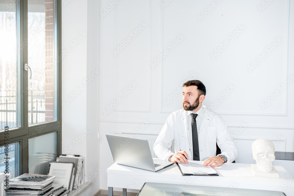 Handsome male doctor in the medical gown working with laptop sitting in the beautiful white office interior