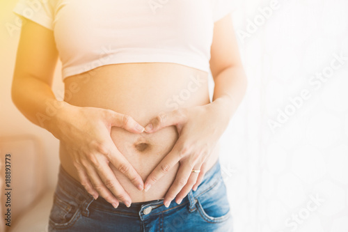 Pregnant Asian Woman holding her hands in a heart shape on her belly near window at home.copy space. © MIA Studio