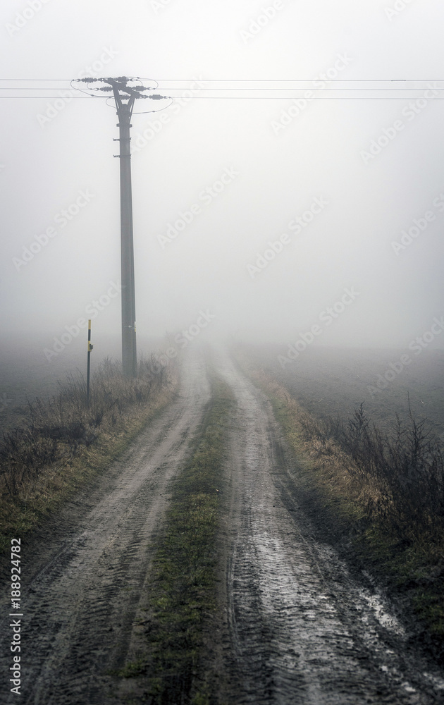 a column with an electric line on a muddy field path emerging from the mist in the highlands