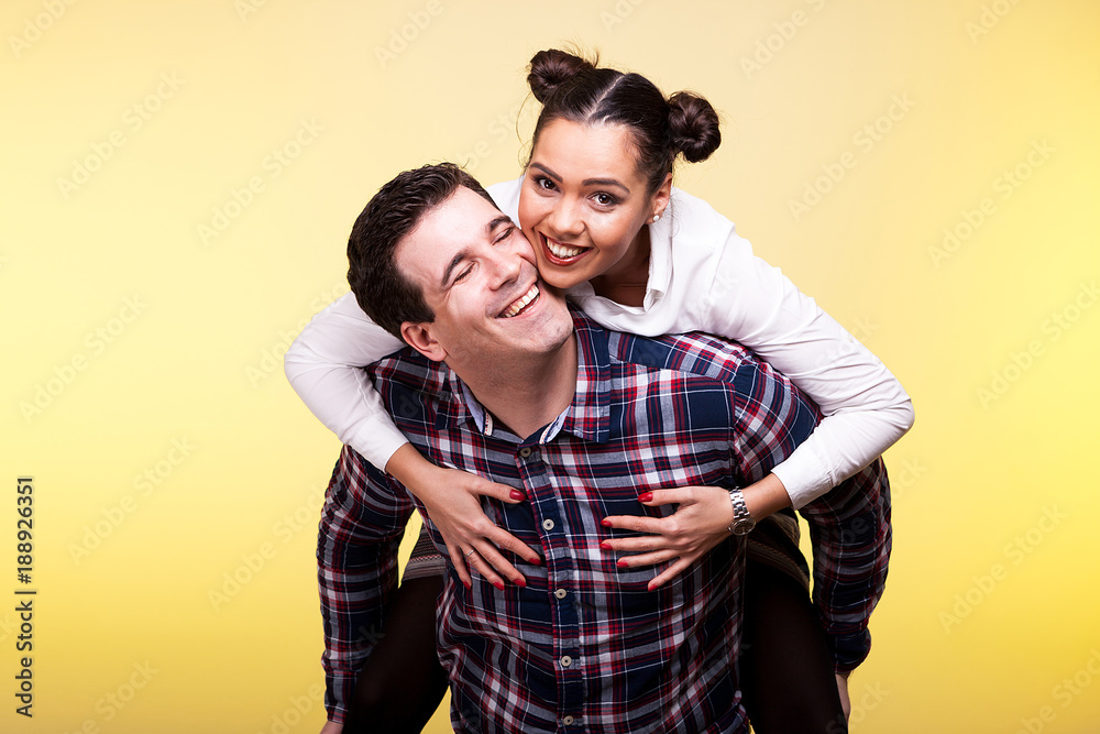 Wife in her husband back looking at the camera and smiling. Yellow background and studio photo
