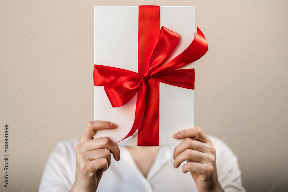 A woman in a white shirt holds a white gift box with a red satin bow and hides her head behind a box A woman in a white shirt holds a white gift box with a red satin bow and hides her head behind a box