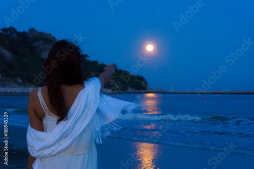 back view beautiful asian woman point finger to full moon over the seascape and mountain with reflection on the water surface at night.