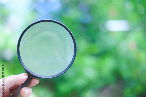 Hand holding a magnifying glass on blurred natural green background