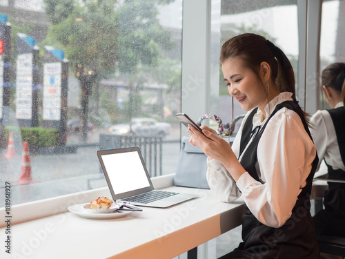 Side view of Asia young business woman in white shirt using smartphone and laptop sitting near window outside raining and uses laptop and smartphone