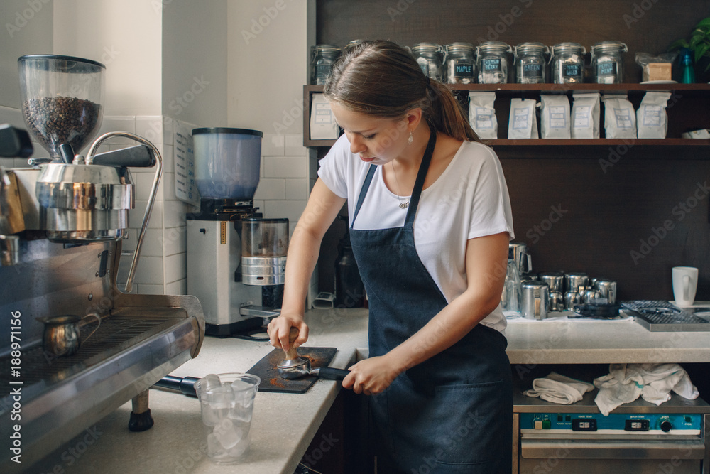 Young Caucasian woman barista using tamper to press ground coffee into portafilter to make espresso hot drink. Small local business work in cafe. Young Caucasian woman barista using tamper to press ground coffee into portafilter to make espresso hot drink. Small local business work in cafe.
