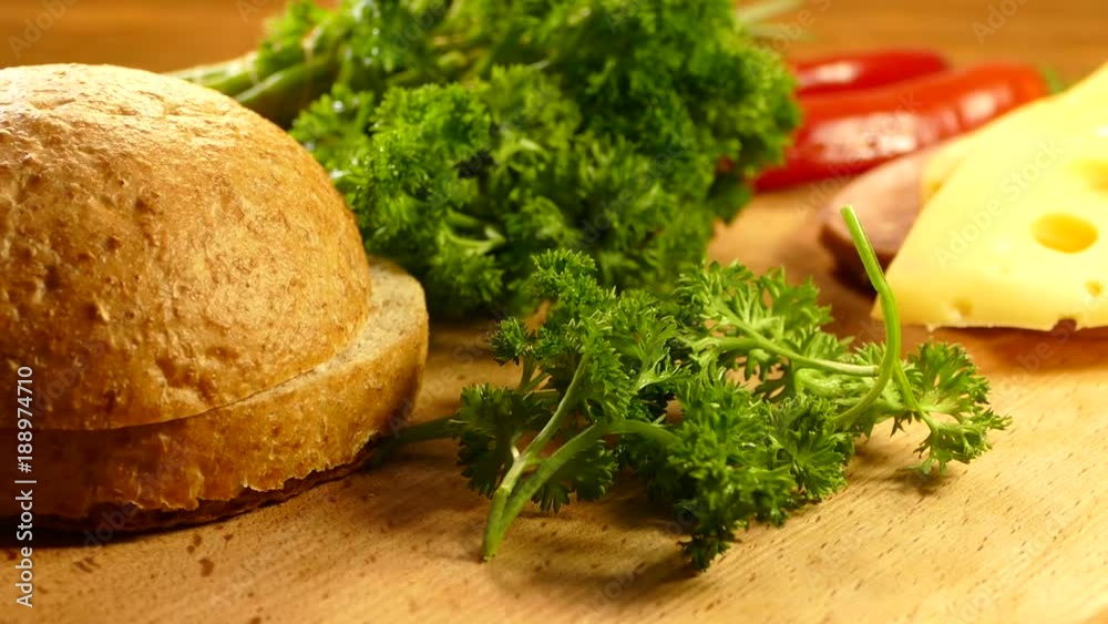 Appetizing sandwich is prepared on a cutting board. Bun, salad, parsley, sausage and cheese are cut into a sandwich. Appetizing sandwich is prepared on a cutting board. Bun, salad, parsley, sausage and cheese are cut into a sandwich.