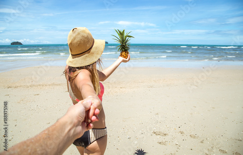 beautiful girl in swimsuit and pineapple walks on the beach holding the hand of the guy