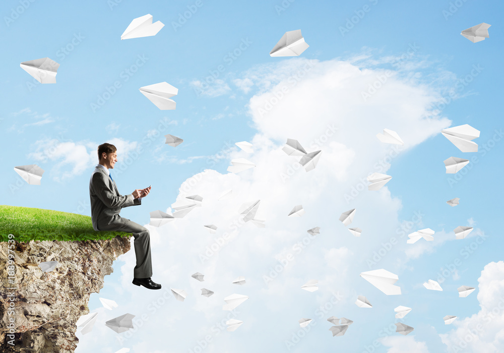 Elegant businessman on rock edge making calls and paper planes flying around Elegant businessman on rock edge making calls and paper planes flying around