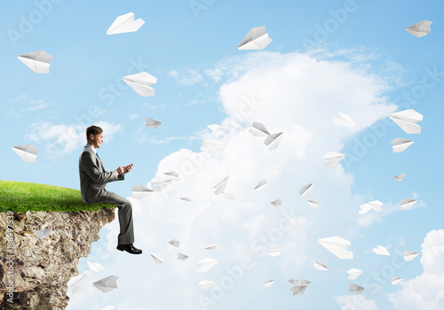 Elegant businessman on rock edge making calls and paper planes flying around