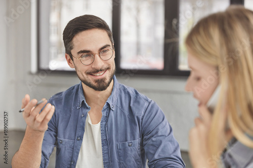 Cheerful bearded handsome male wears round glasses, has pleasant conversation with female colleague, try to discuss main issues of their work. Pretty young woman talks via modern cell phone. photo