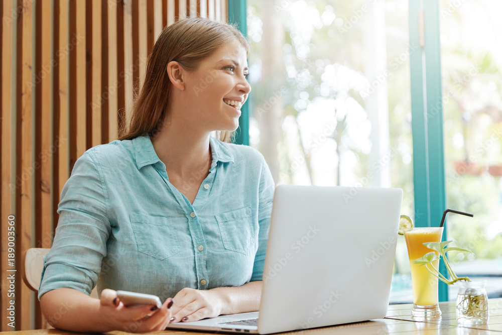 Cheerful attractive female copywriter earns money online, works remotely at laptop computer, holds modern cell phone as waits for important call, sits in coffee shop, looks out of big window Cheerful attractive female copywriter earns money online, works remotely at laptop computer, holds modern cell phone as waits for important call, sits in coffee shop, looks out of big window