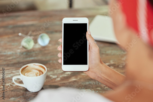 Unrecognizable woman reads news in social networks on mobile phone while sits at coffee shop. Female`s hand holds modern cell phone with blank copy screen for your advertising text messagge. photo