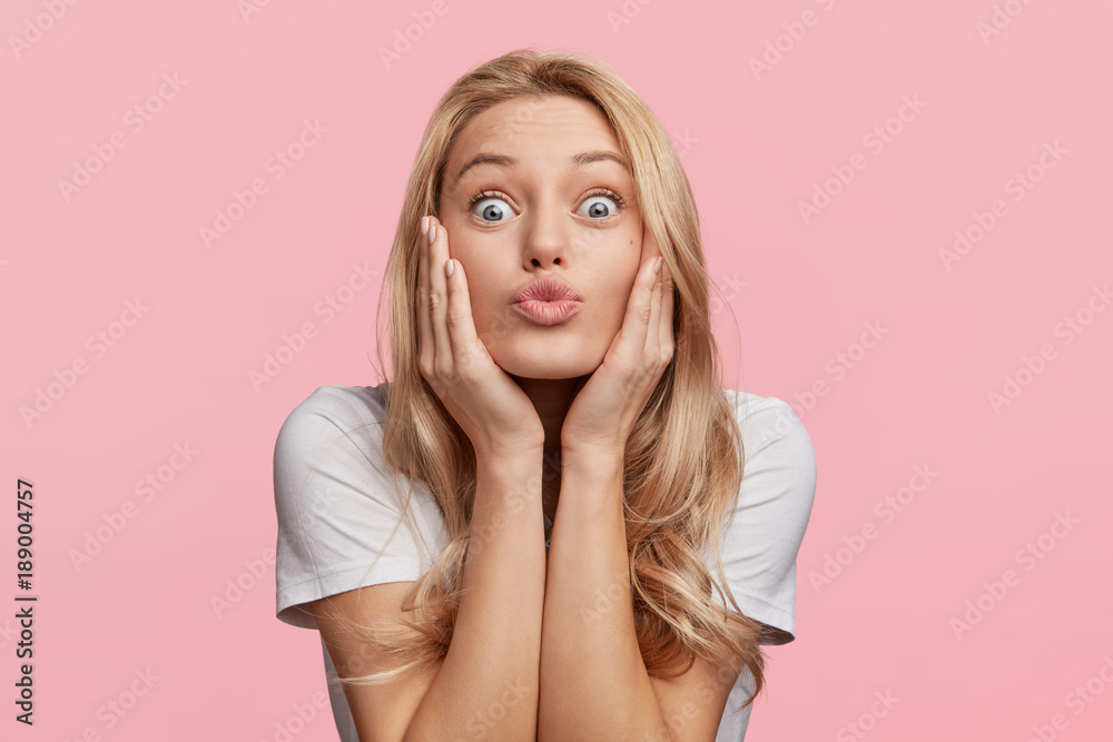 Pleased funny woman pouts lips and looks with bugged eyes at camera, makes  grimace, has attractive look, isolated over pink background. Indoor shot of  beautiful young female poses indoor in studio Stock