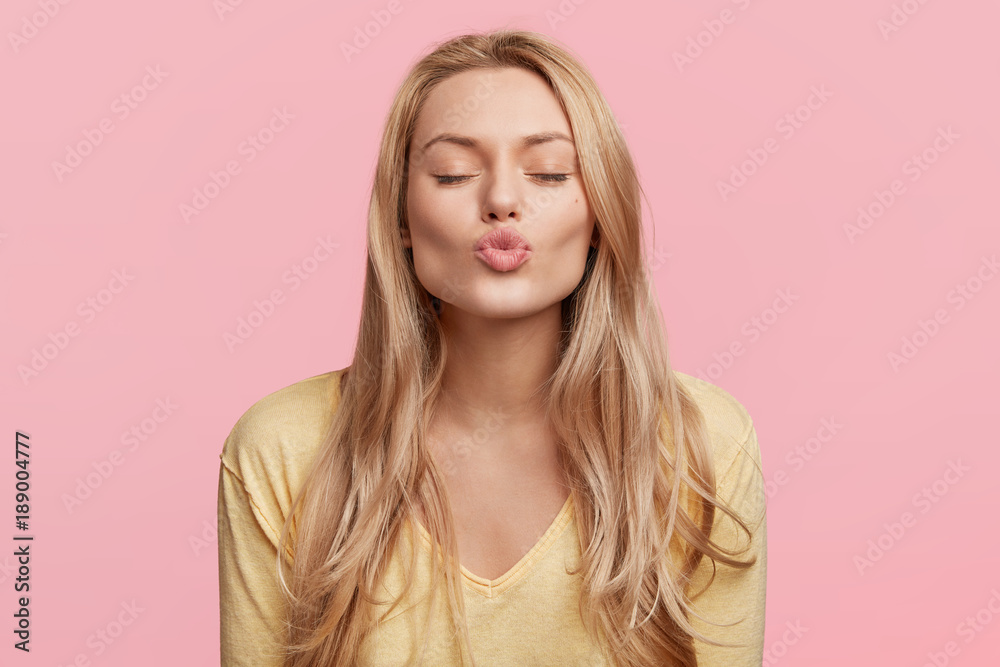 Fototapeta premium Lovely young woman with rounded lips and shut eyes, makes kiss, has long blonde hair, poses against pink background, being pleased, expresses her positive feeling, imagines something pleasant