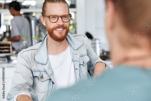 Best friends meet together at cafe, share news and being glad to see each other. Cropped shot of hipster male with thick red beard communicates with unrecognizable man, discuss future plans.