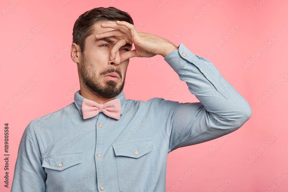 Displeased brunet male plugs nose as smells something stink and unpleasant, feels aversion, isolated over pink background. Young attractive man hates disgusting scent which comes from next room
