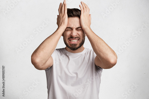 Young European bearded young male clenches teeth in desperation, regrets about something, dressed in casual t shirt, isolated over white background. Negative human emotions and feelings concept photo