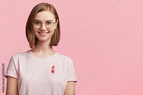 Cheerful female volunteer involved in helping people to cure disease, wears pink ribbon which symbolizes struggle from women`s disease mammary cancer, poses against pink background with copy space