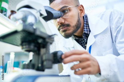 Portrait of young Middle-Eastern scientist looking in microscope while working on medical research in science laboratory, copy space