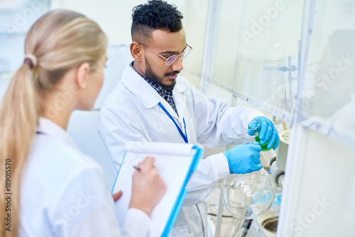 Portrait of two modern scientists working in medical laboratory doing research, focus on Middle-Eastern man pouring colored liquid to test tubes