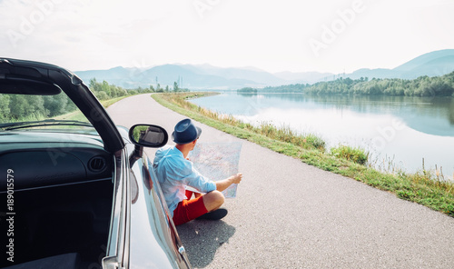 Man reads roads map sitting near his cabriolet on picturesque mountain road