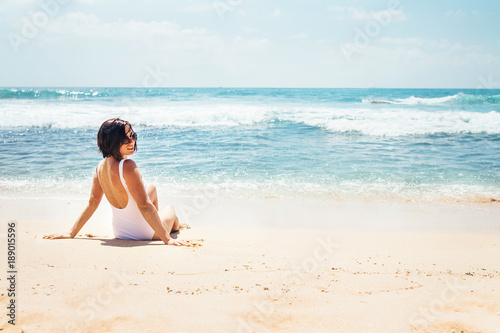 Woman in white swimsuit sits on empty ocean beach in sunny day