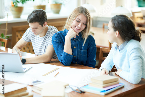 Cheerful blond-haired student with charming smile chatting animatedly with her friend while sitting at wooden table of library, their handsome groupmate wrapped up in doing homework