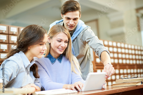 Cheerful group of university students watching funny video on digital tablet while distracted from doing homework in spacious reading room