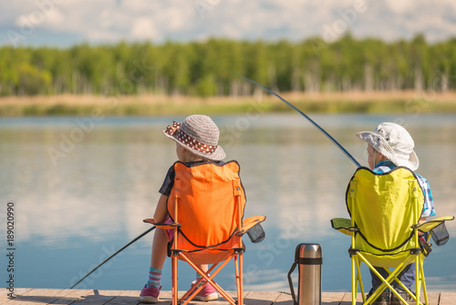 children with fishing rods sit on a wooden pier and fish at the lake