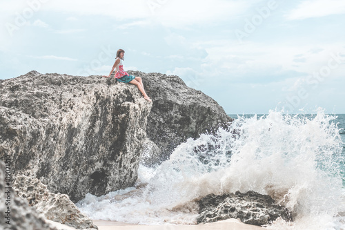 Beautiful young woman sitting on the stone on the seashore. Tropical island of Bali, Indonesia.