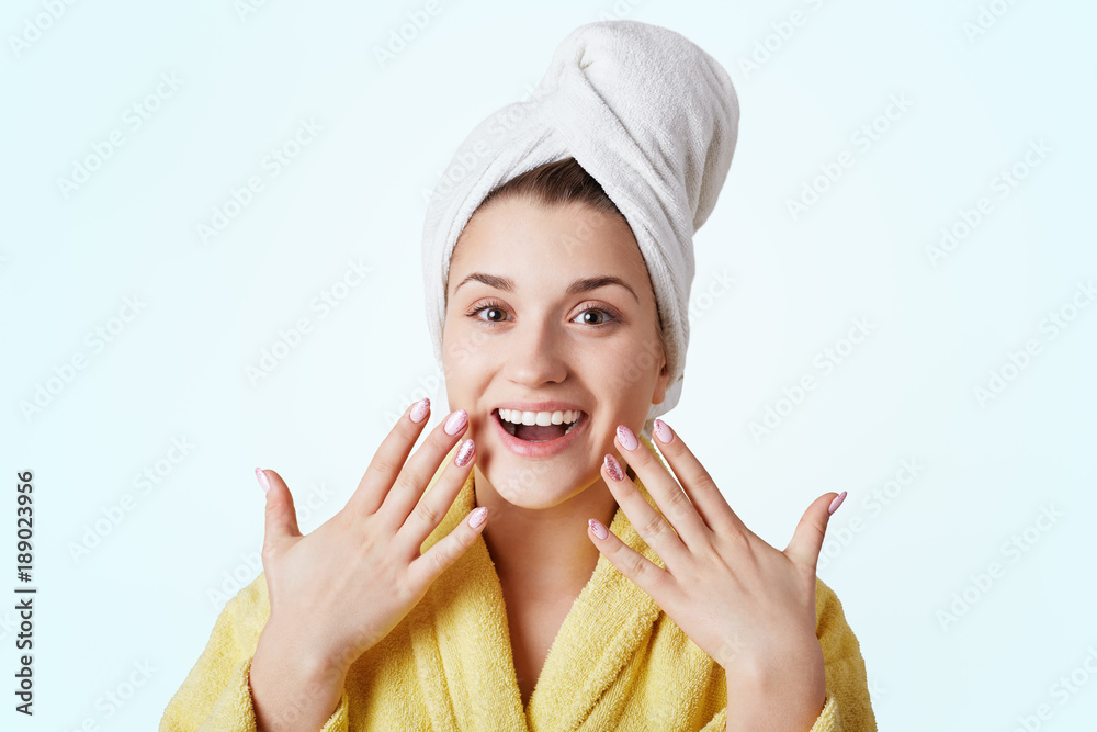 Excited happy female takes shower, demosntrates pure healthy skin and beautiful manicure, takes care of her beauty and appearance, wears bathrobe and towel on head, isolated on white background Excited happy female takes shower, demosntrates pure healthy skin and beautiful manicure, takes care of her beauty and appearance, wears bathrobe and towel on head, isolated on white background