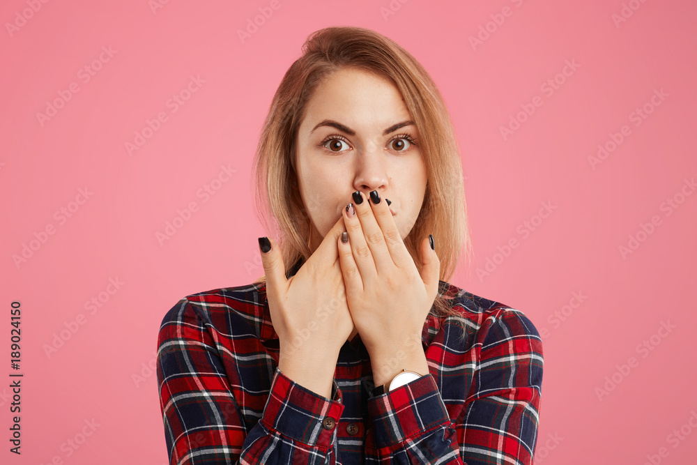 Lovely young female with manicure covers mouth with puzzled expression, tries to keeps silence, not gossip and tell confidential information to anybody, poses against pink studio background. Lovely young female with manicure covers mouth with puzzled expression, tries to keeps silence, not gossip and tell confidential information to anybody, poses against pink studio background.