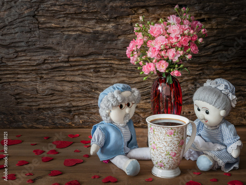 Still life visual art of grandparent dools sitting with a cup of coffee and pink rose in red vase with paper hearts falled around on wooden floor with wooden slab background photo