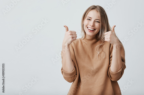 Portrait of fair-haired beautiful female student or customer with broad smile, looking at the camera with happy expression, showing thumbs-up with both hands, achieving study goals. Body language photo