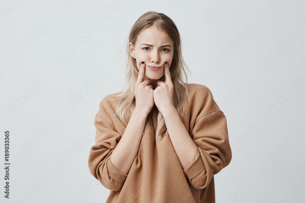 Studio shot of beautiful blonde woman making grimace, touching her cheeks with fingers, making herself smile, frowning face, being displeased and upset. Face expression and negative emotions