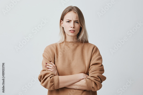 Waist-up portrait of beautiful girl with blonde straight hair frowning her face in displeasure, wearing loose long-sleeved sweater, keeping arms folded. Attractive young woman in closed posture. photo