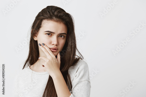 Pretty brunette girl with dark eyes wearing hair loose frowning her eyebrow, looking at camera with placid and thoughtful look. Pensive young female with puzzled expression thinking something over
