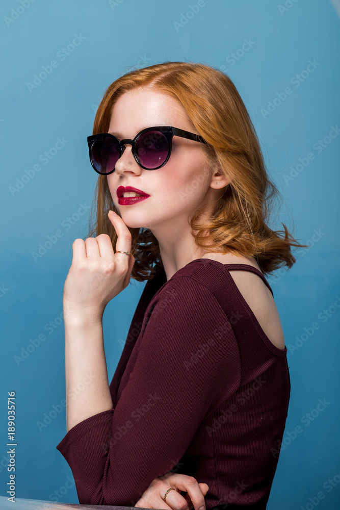 Redhead sexy beautiful girl with makeup and red lips in sunglasses on blue background.