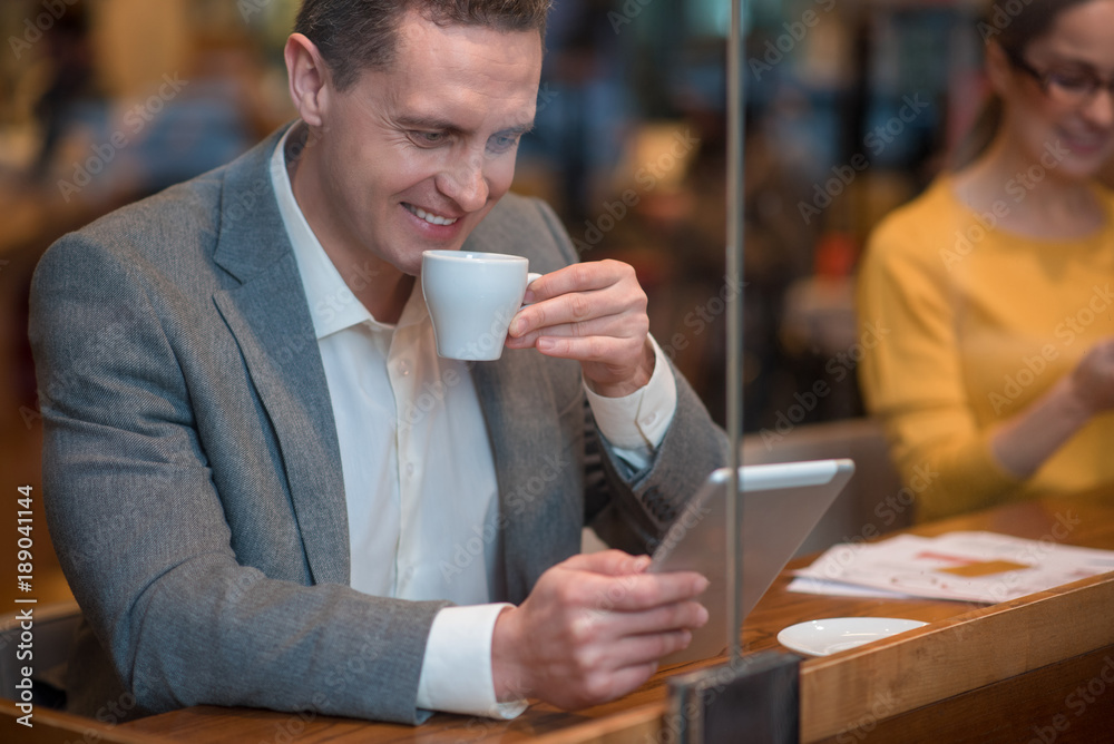 Outgoing man drinking cup of appetizing coffee while looking at digital device. He sitting near smiling female at table in cafe. Rest concept