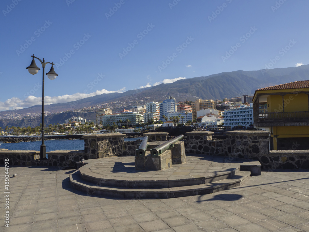 Fototapeta premium old copper cannons on paved sea front with lamp and view on sea with big hotel resort buildings panorama of Puerto de la cruz, tenerife with green hill and blue sky background