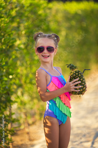 Little girl in a swimsuit and sunglasses on the beach with a cocktail in pineapple
