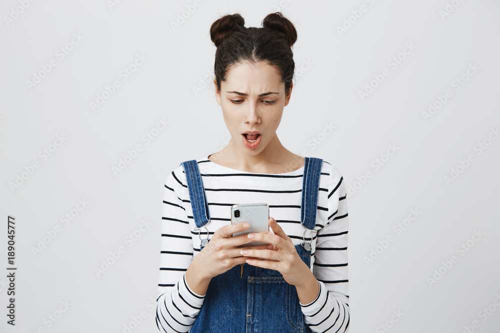 Stunned brunette young woman with hair knots in denim overall, received message on smartphone with bad news, being shocked and frowning face in displeasure, isolated against gray wall.